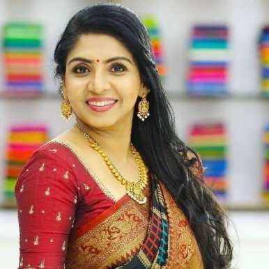 Priyadarshini is smiling, standing, has black hair and a bindi wearing a large dangling earring, gold necklace and a red printed saree.