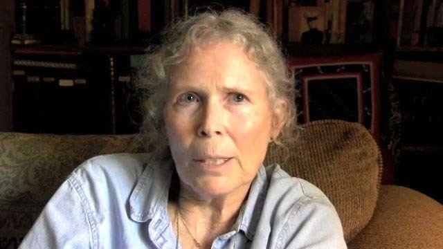 Prudence Farrow sitting on the couch while wearing a light blue blouse