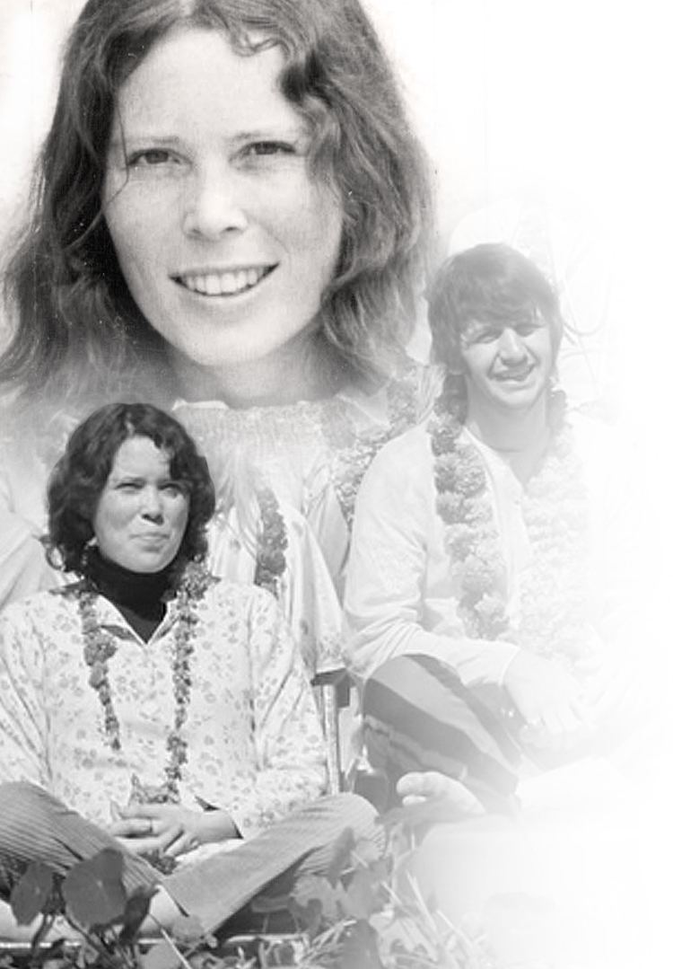 Prudence Farrow smiling while sitting on the floor and wearing a floral blouse and garland