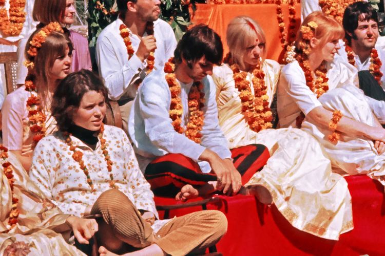 Prudence Farrow sitting on the floor with other people and wearing a floral blouse and garland