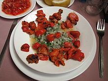 On top of a brown table, there is a white plate of cooked Pyura chilensis and greens, along with a plate of red sauce and a plate of lemon at the top, a glass of water and a silver fork at the right.