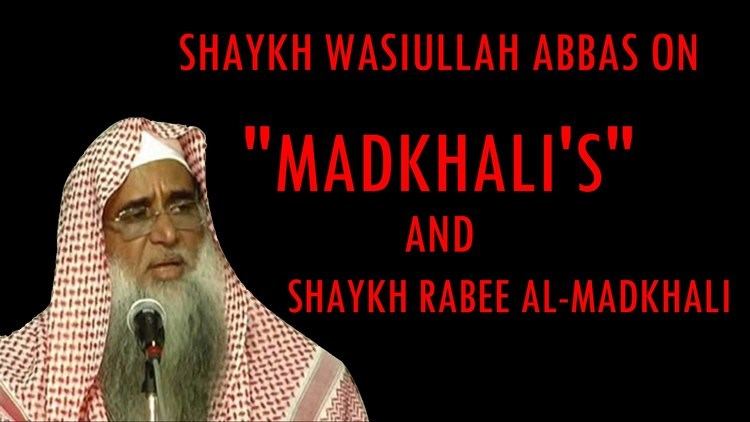 Rabee al-Madkhali speaking while wearing white and red keffiyeh and eyeglasses