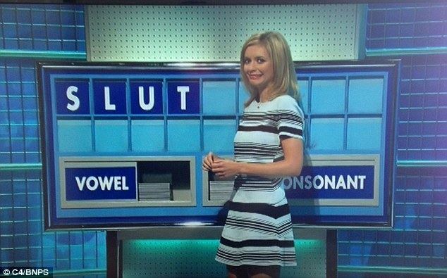 On Countdown, Rachel Riley is smiling awkwardly at the UK game show, both hands holding each other, a background of blue tiles, with a board at the back with the word “SLUT” at the top and a vowel on the lower left with piled tile letters, and a consonant on the lower right with piled tile letters, she is wearing a blue dress with black and white lines.