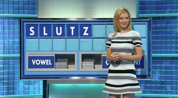 On Countdown, Rachel Riley is smiling, has blonde hair, both hands holding each other, a background of blue tiles, with a board at the back with the word “SLUTZ” at the top and a vowel on the lower left with piled tile letters, she is wearing a blue dress with black and white lines.
