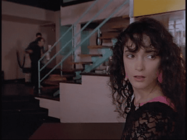 Rainbow Harvest with black curly hair while wearing pink earrings and black lace blouse in a scene from the 1984 tv series Miami Vice