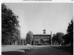 A Black and White front view of the Rainhill Hospital with visible lampposts and trees along the pathway.