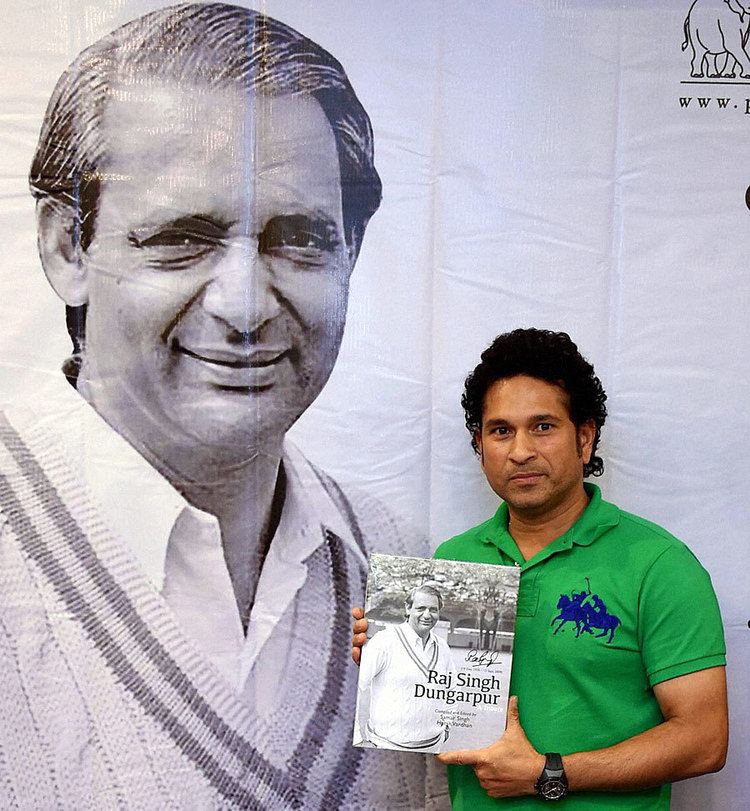 Sachin Tendulkar smiling and standing while holding a book, Raj Singh Dungarpur - A Tribute, in Mumbai while an image of Raj Singh Dungarpur is on a banner in the background, during the book launching on July 25, 2014. Sachin with black curly hair, a black wristwatch, wearing a blue embroidered logo of two Polo players on their horses on a green collared shirt. While Raj on the banner is wearing a white collared shirt under a knitted vest.