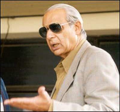 Raj Singh Dungarpur talking to someone with an open palm gesture, a former BCCI President, who has white hair, vintage sunglasses on, and wearing a caramel collared shirt under a beige coat.