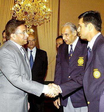 Raj Singh Dungarpur looking at the Pakistan President, Pervez Musharraf (on the left) while shaking hands with Indian Cricket captain, Rahul Dravid (on the right) standing beside him with some men in suits and tie in the background. Perez has brown hair, eyeglasses, finger ring, wearing a gray suit and tie and a white collared inner shirt. Raj in white hair, wearing a pair of light-gray slacks, a white collared shirt, a navy-blue printed necktie, and a BCCI logo patched on the left of his midnight-blue coat. And Rahul is wearing a white collared shirt, a navy-blue printed necktie, and a BCCI logo patched on the left of his midnight-blue coat.