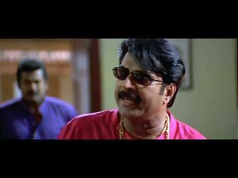 Mammootty with a mustache, wearing sunglasses, gold necklaces, and a pink shirt with a man on his back wearing a purple shirt in a movie scene from Rajamanikyam (2005 film).