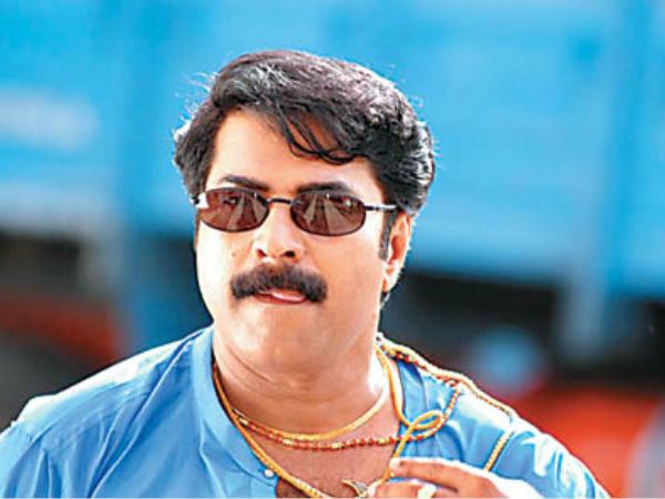 Mammootty with a serious face and mustache, with his tongue out, wearing sunglasses, gold necklaces, and a blue shirt in a movie scene from Rajamanikyam (2005 film).