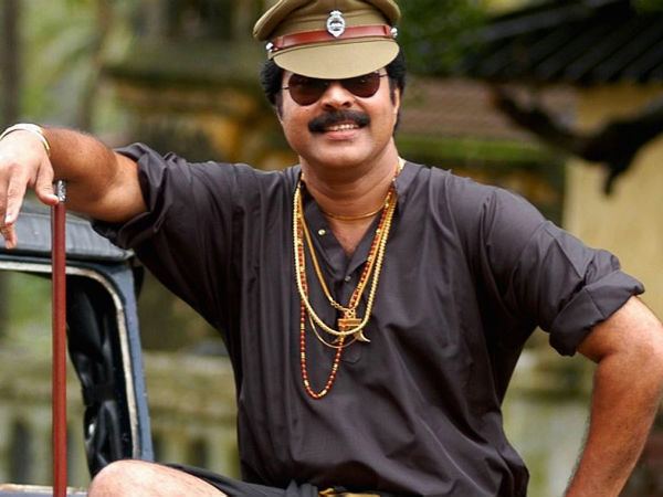 Mammootty with a smiling face while his hand on his waist, wearing sunglasses, a green hat, gold necklaces, a black polo shirt, with a golden stripped black lungi in a movie scene from Rajamanikyam (2005 film).