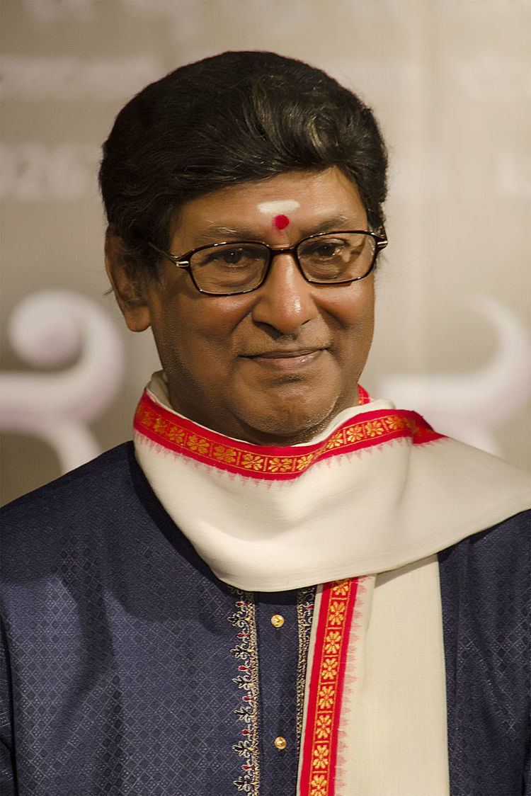 Rajesh smiling while looking at something, with a white & red tika on his forehead, and wearing eyeglasses, Indian attire blue kurta, and a white and red scarf with yellow flowers on the side