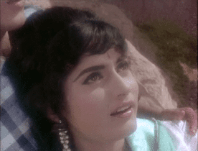 Rajshree looking above, with a smiling face, wearing earrings and an avocado green top with a man holding her shoulder wearing a blue checkered shirt.