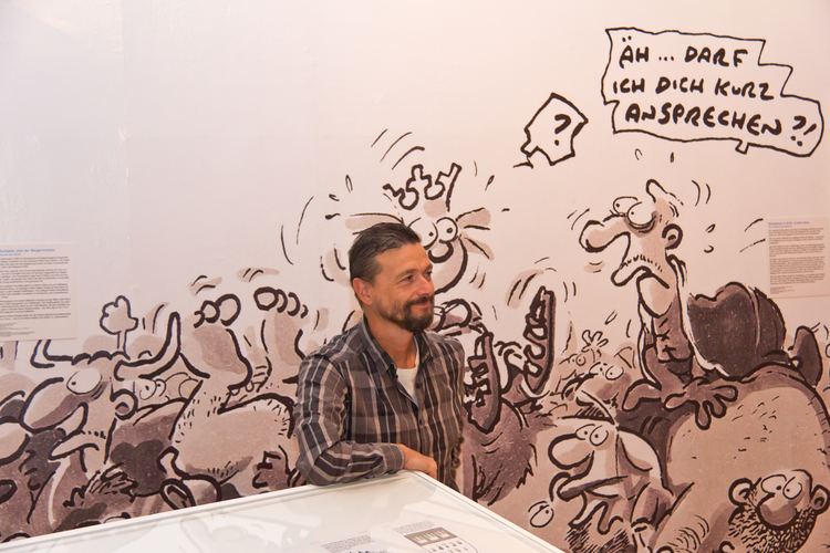 Ralf König with a tight-lipped smile with a background of comic drawings, with beard and mustache, and wearing a gray checkered long sleeve over a white shirt.