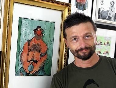 Ralf König with a serious face while leaning on a frame with his drawing on it, with beard and mustache, and wearing a gray shirt.