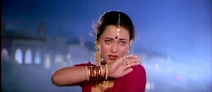 In the movie scene of  Ram Teri Ganga Maili 1985, Mandakini is serious, standing in the middle with Indian architecture buildings behind her, has black hair and a bindi on her forehead, wearing a maang tikka on her forehead, long gold earrings, gold necklace, red and gold bracelets, rings on her right hand, and a red dress.