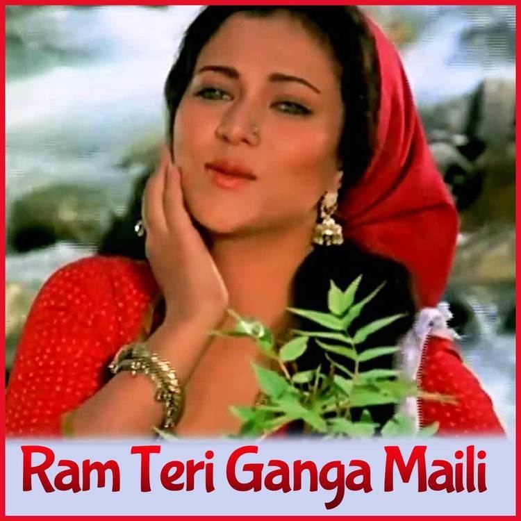 In the movie scene of  Ram Teri Ganga Maili 1985, Mandakini is smiling, standing in the middle with leaves in front and a river behind her, right hand under her right jaw,  has black hair, brown eyes, wearing a nose piercing, gold earrings, gold ring, necklace, gold bracelets in her right hand, red cloth over her head and red long sleeve dress with white dots.