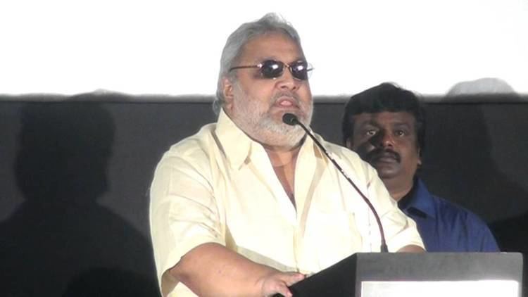 Ramkumar Ganesan wearing sunglasses, a yellow polo shirt, standing on a podium with a microphone with a man on his back wearing a blue shirt.