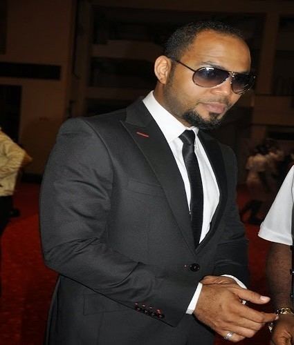 Ramsey Nouah wearing a black coat, white long sleeves, a black necktie, and shades