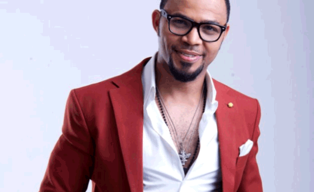Ramsey Nouah smiling while wearing a red coat, white long sleeves, necklace, and eyeglasses