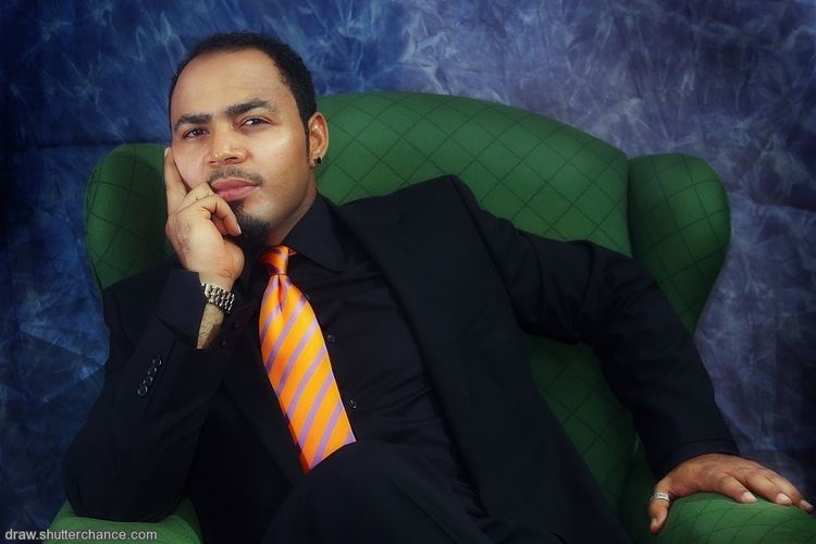 Ramsey Nouah sitting on a green chair while wearing a black coat, black long sleeves, and striped necktie