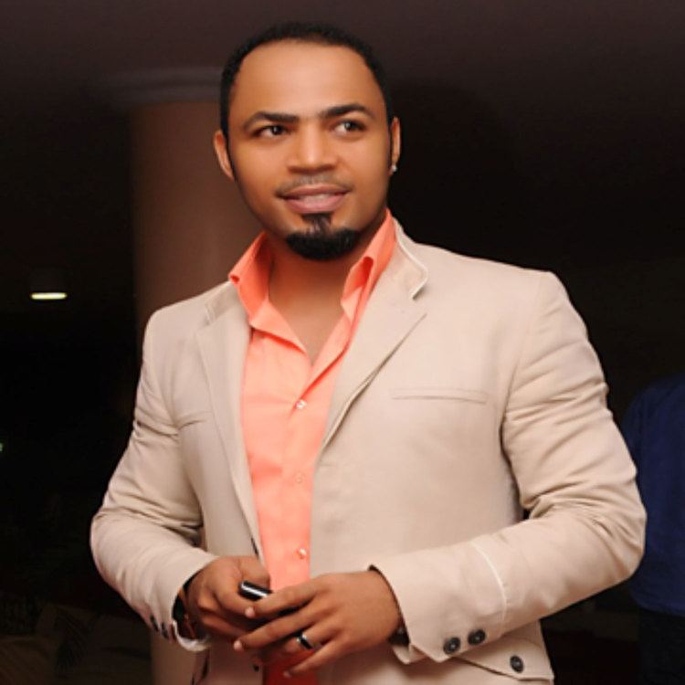 Ramsey Nouah smiling and wearing a beige coat and orange long sleeves