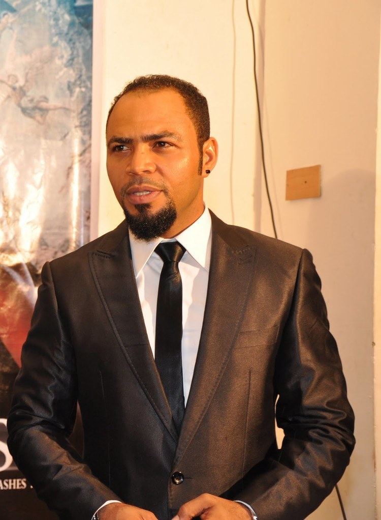 Ramsey Nouah wearing a black coat, white long sleeves, and a black necktie