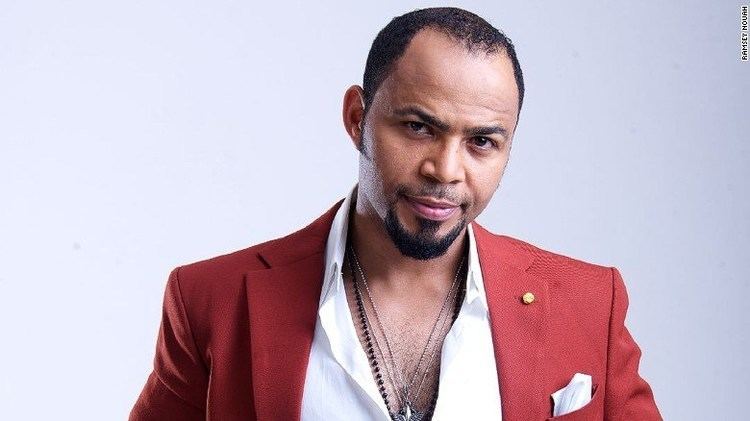 Ramsey Nouah wearing a red coat, white long sleeves, and necklace