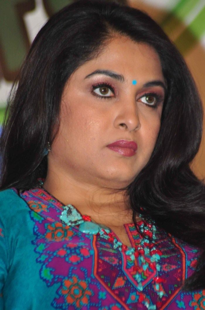 Ramya Krishnan looking afar with a poker face while wearing a blue-green, purple, and orange dress and blue earrings