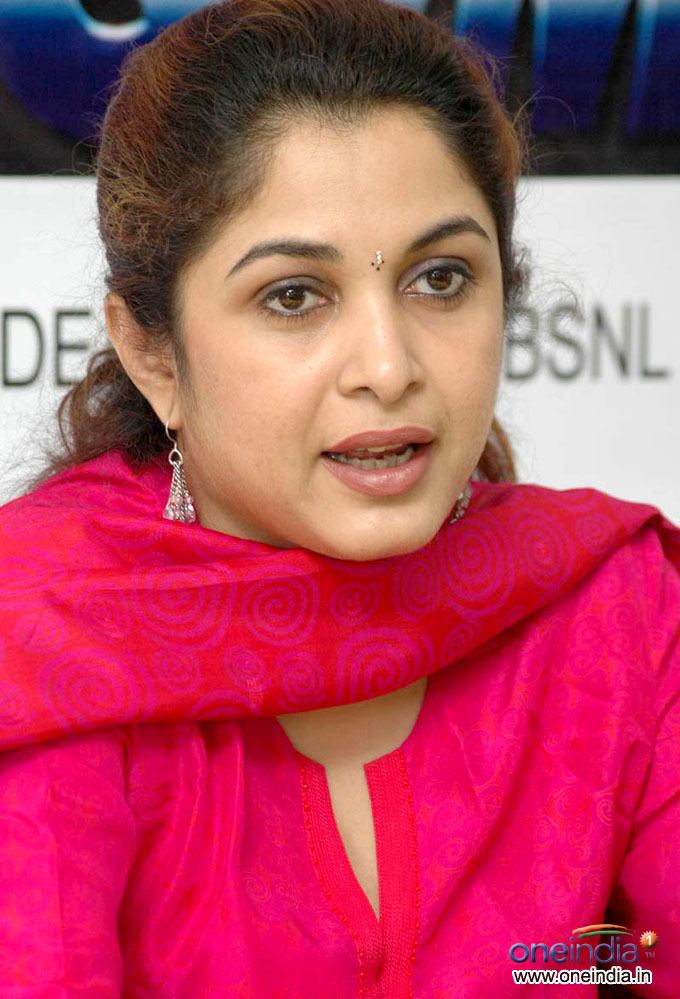 Ramya Krishnan talking to someone while wearing a red dress, red scarf, and earrings