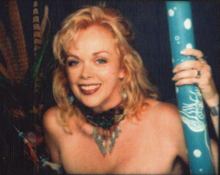 Raven De La Croix smiling while holding a wrapping paper roll and wearing a necklace, rings, and earrings