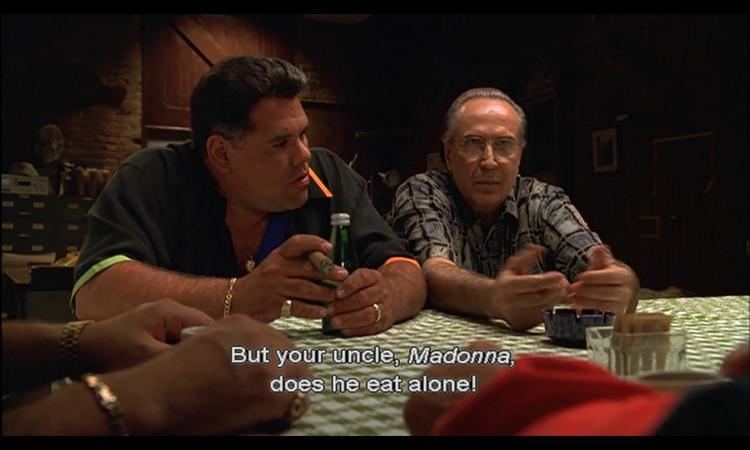 Ray Curto talking to a man while wearing eyeglasses and a gray and black polo in a scene from the 1999 tv series The Sopranos