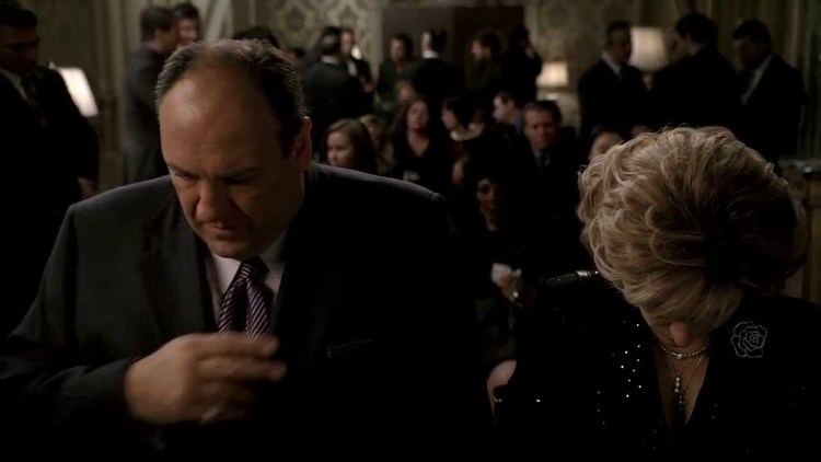 A man standing beside the woman while wearing a black coat, long sleeves, and a necktie in a scene from the 1999 tv series The Sopranos