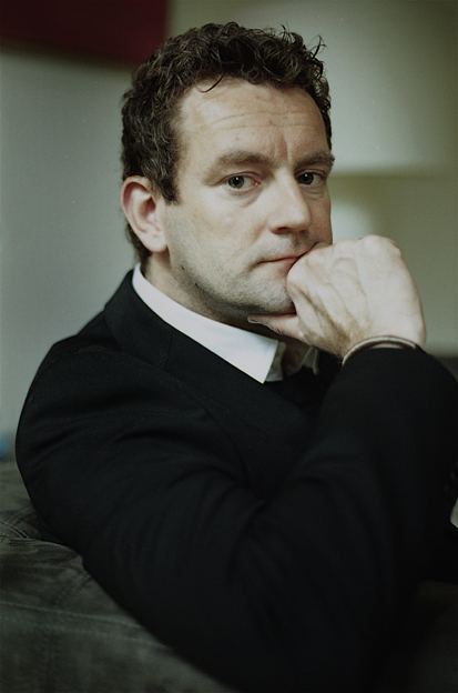 Ray Kluun with a serious face while sitting on a couch and his hand on his chin, with curly hair, and wearing a black coat over white long sleeves.