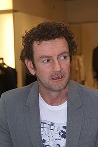Ray Kluun with a serious face, curly hair, wearing a gray coat over a white shirt with black print.