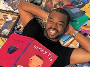 LeVar Burton is smiling while lying on books and a book named Enemy Pie on his chest, with a beard and mustache, wearing a watch, an earring, and a black shirt.