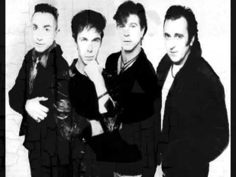 Members of the Real Life band, Alan Johnson, David Sterry, Richard Zatorski, and Danny Simcic with serious faces and wearing black jackets.