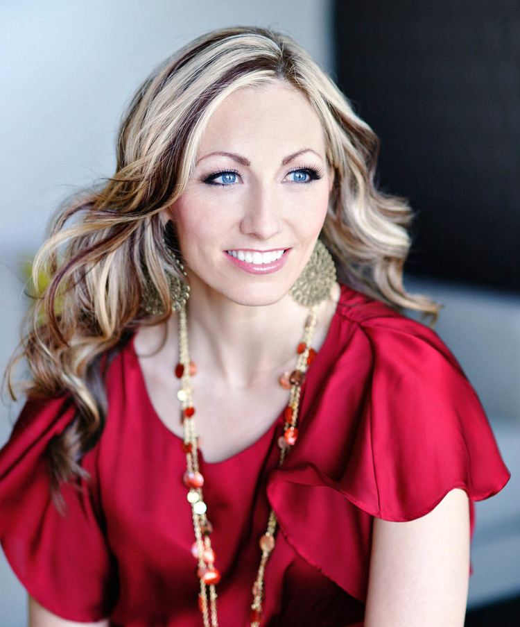 Rebecca Musser smiling with her wavy blonde highlights and wearing dangled earrings, a long necklace, and red blouse
