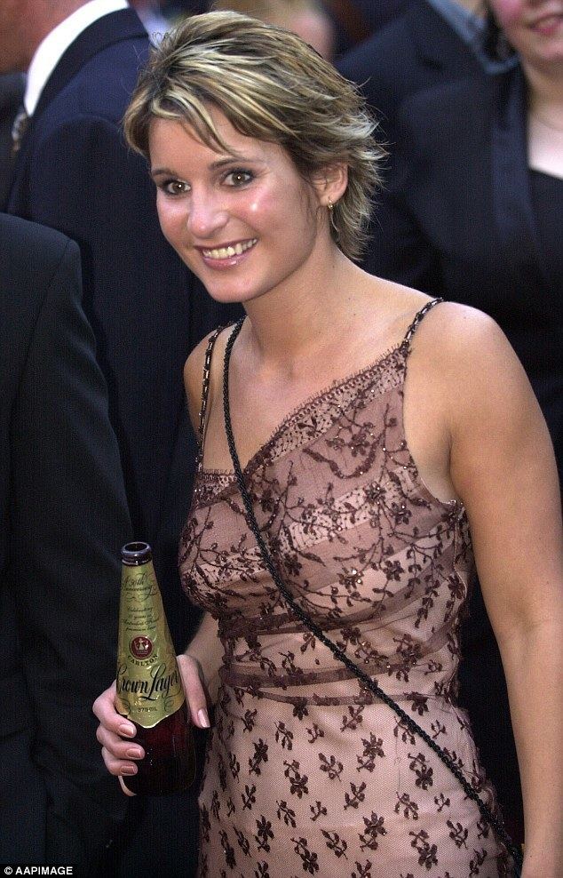 Regina Bird is smiling, and has black-blonde hair, holding a bottle of beer in her right hand, wearing gold earrings, a black sling bag, and a brown dress with a flower design.