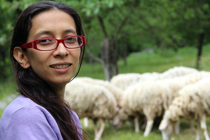 Regina José Galindo smiling, having long black hair, wearing red eyeglasses and a purple shirt with sheep in her background