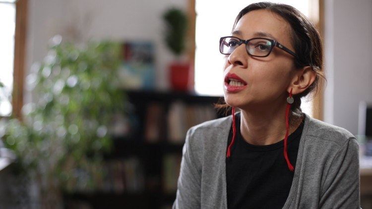 Regina José Galindo is talking during an interview, wearing eyeglasses, red earrings, and a gray blazer over a black shirt.