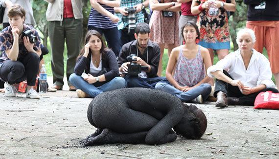 Regina José Galindo's body was entirely covered in coal, curled on the hard ground, crouched down, and rounded out her back, her knees, and elbows hugging against her ribs while surrounded by people watching her.