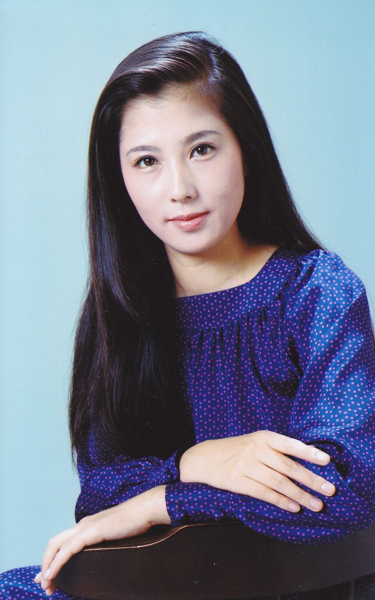 Reiko Ohara smiling while sitting on the chair and wearing a blue dress