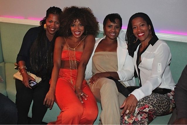 Reina King (4th, from the left) and her friends are smiling while sitting on a light green couch. Reina is wearing loop earrings, a white and black long sleeve top, and animal-print pants with a floral design.