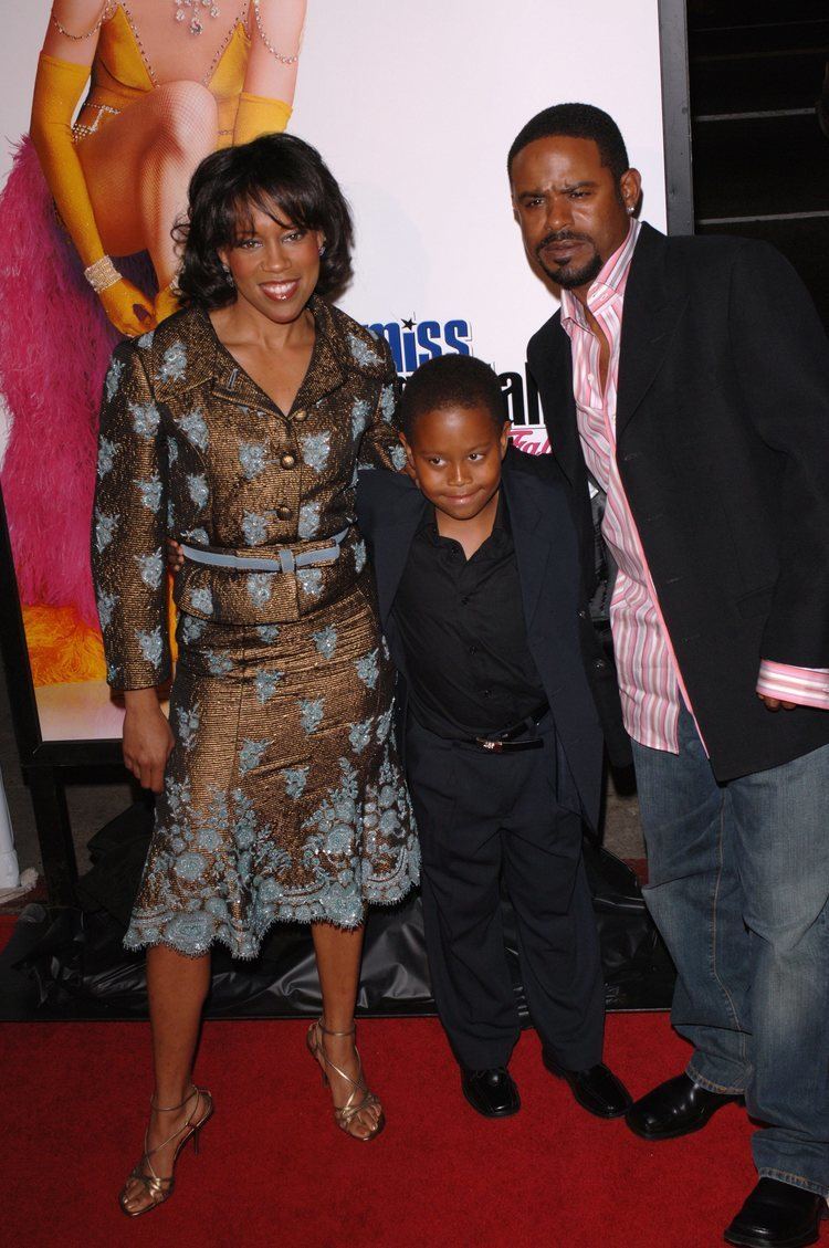 Regina King, Ian Alexander, Jr., and Ian Alexander Sr. (from left to right) at the US premiere of her new movie Miss Congeniality 2. Regina with wavy hair, wearing a brown and gray dress, and brown sandals, Ian Jr. is wearing a dark blue coat over a black polo shirt, black belt, black pants, and black shoes while Ian Sr. with beard and mustache, wearing earrings, a black coat over a white and pink striped long sleeves, blue jeans, and black shoes.