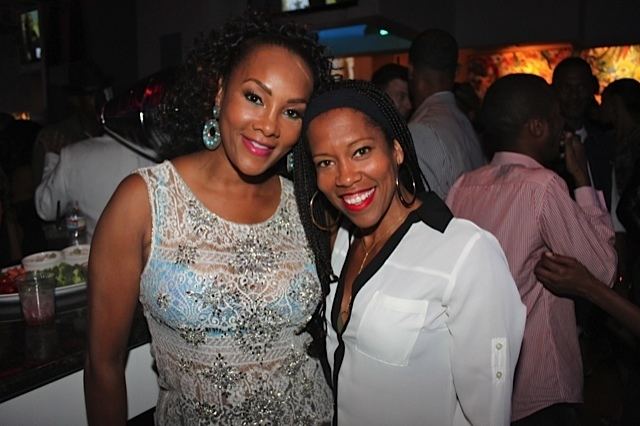 Reina King and Vivica Fox are smiling during Vivica’s 49th birthday bash. Reina is wearing a black headband, loop earrings, and a white and black long sleeve blouse while Vivica is with curly black hair, wearing blue earrings and a white see-through sleeveless top over a blue bra.
