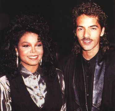 Janet Jackson and René Elizondo Jr. are smiling while Janet with black curly hair is wearing hoop earrings and a brown long sleeve blouse under a black vest. René with a mustache and curly hair is wearing a black long sleeve under a dark violet coat