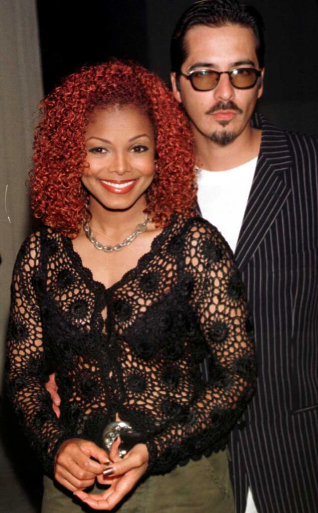 Janet Jackson smiling and René Elizondo Jr. is looking seriously with a mustache and beard. Janet with red curly hair is wearing moss green pants, a necklace, earrings, and a black brassiere under a black long sleeve crochet blouse. René is wearing sunglasses and a white shirt under a blue striped coat