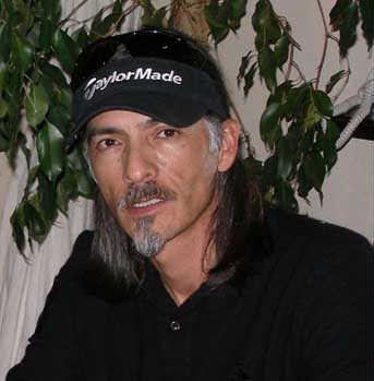 René Elizondo Jr. with a serious face, mustache, beard, shoulder-length hair, and plants in the background while wearing a black polo shirt and a black cap with sunglasses on the top of it